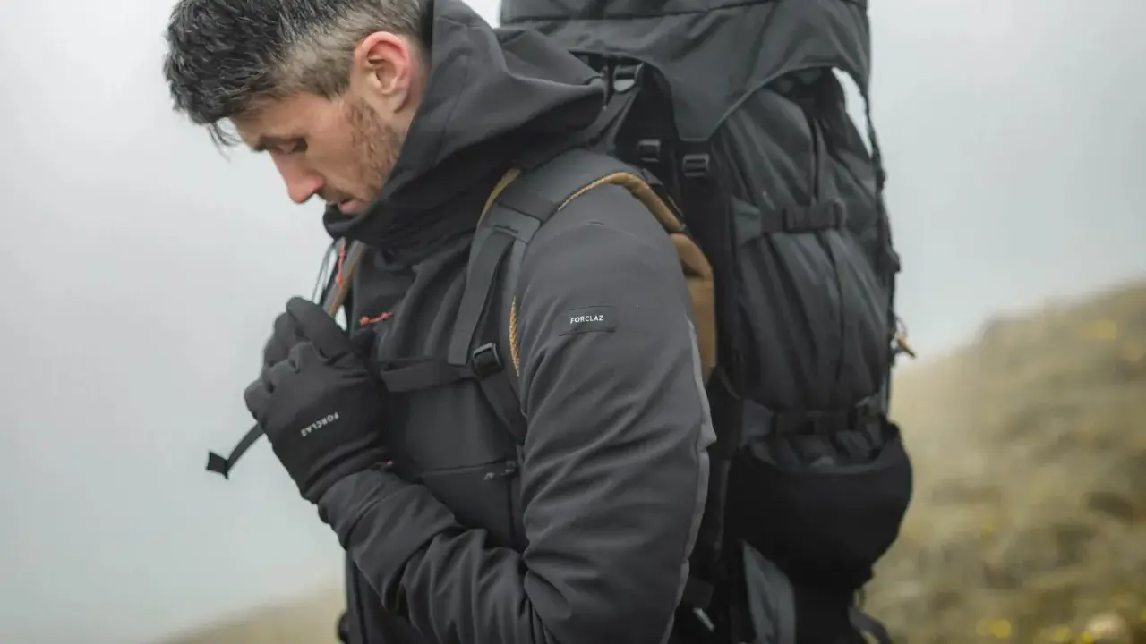 Mężczyzna w ciemnej kurtce softshell i rękawiczkach, z dużym plecakiem, przygotowuje się do wędrówki. Czy softshell jest nieprzemakalny? Ta kurtka wygląda na gotową na deszcz.