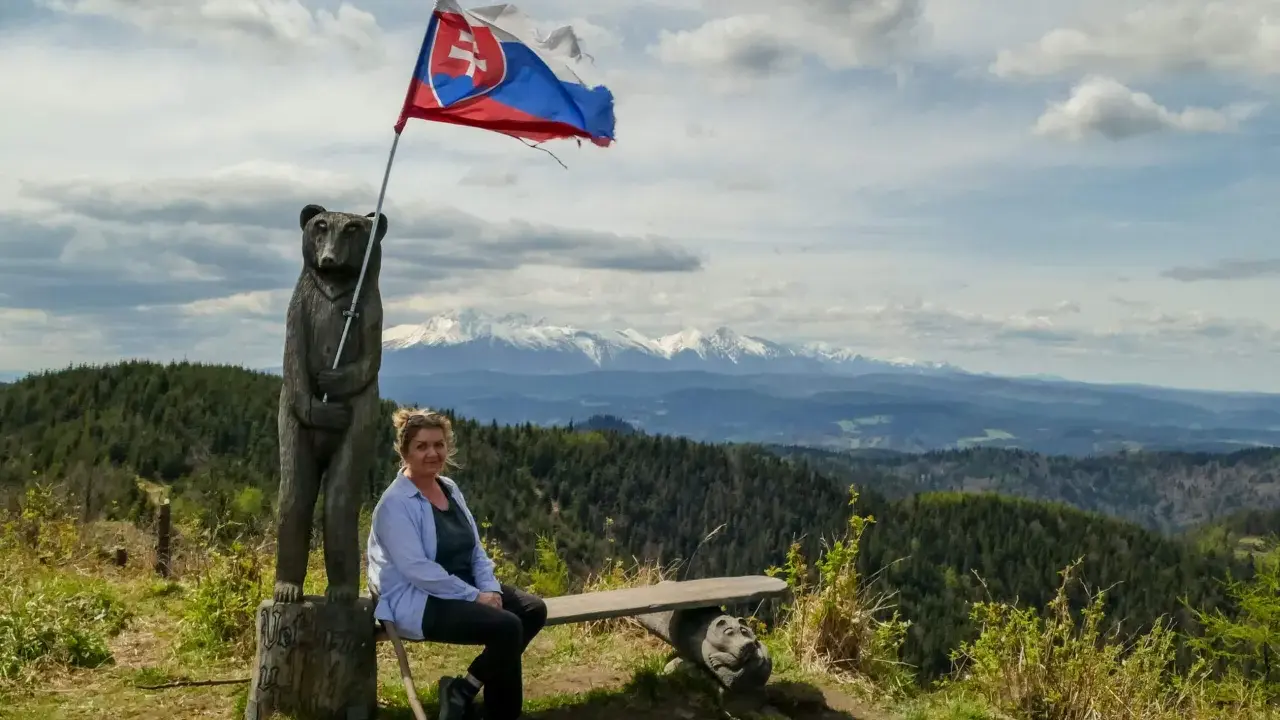 Kobieta siedzi na ławce pod słowacką flagą na wietrznym wierchu. W tle rozciągają się góry i lasy.