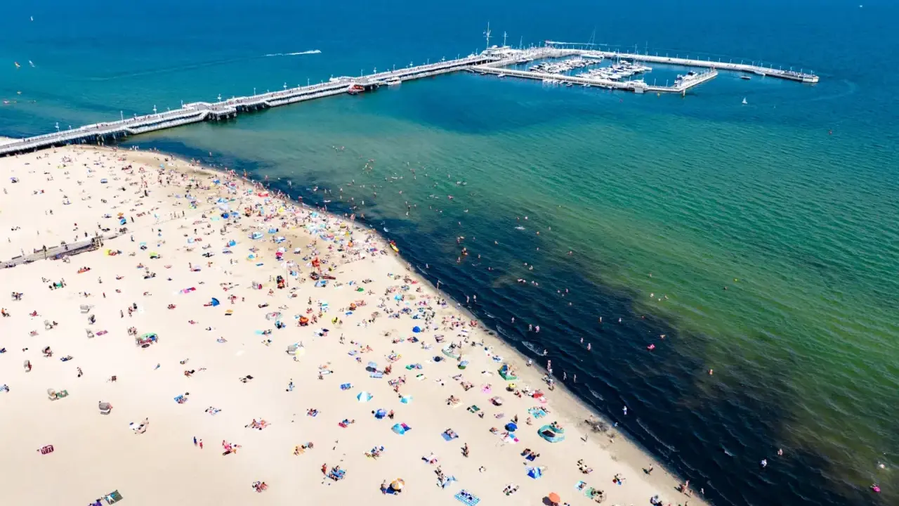 Tłum ludzi wypoczywa na piaszczystej plaży w Sopocie. W oddali widać molo i przystań jachtową.