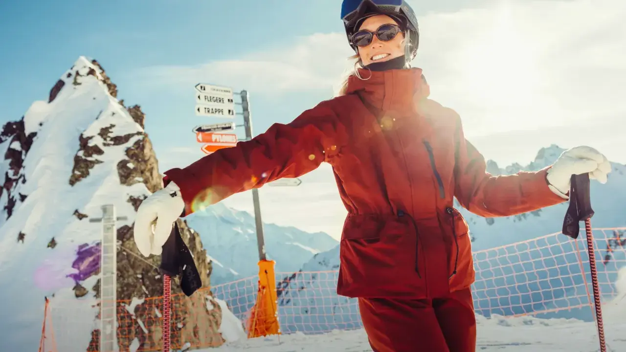 Narciarka w pomarańczowej kurtce Helly Hansen, z nartami i kijami, na tle ośnieżonych gór.