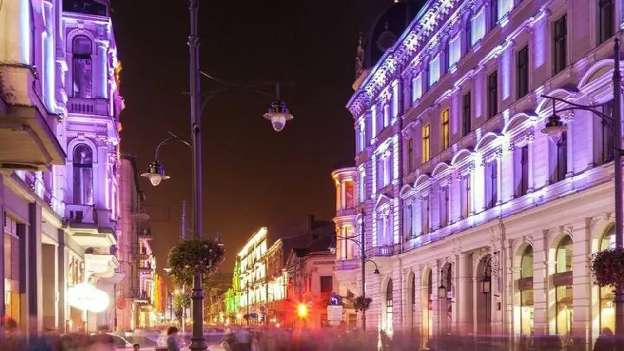 Nocą ulica Piotrkowska w Łodzi rozświetlona fioletowymi i zielonymi światłami. Widać ludzi spacerujących i siedzących w ogródkach kawiarnianych. Co ciekawego w Łodzi? Ta iluminacja!