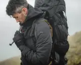 Mężczyzna w ciemnej kurtce softshell i rękawiczkach, z dużym plecakiem, przygotowuje się do wędrówki. Czy softshell jest nieprzemakalny? Ta kurtka wygląda na gotową na deszcz.