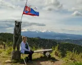 Kobieta siedzi na ławce pod słowacką flagą na wietrznym wierchu. W tle rozciągają się góry i lasy.