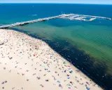 Tłum ludzi wypoczywa na piaszczystej plaży w Sopocie. W oddali widać molo i przystań jachtową.