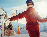 Narciarka w pomarańczowej kurtce Helly Hansen, z nartami i kijami, na tle ośnieżonych gór.