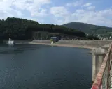 Betonowa zapora na jeziorze żywieckim, otoczona zielonymi wzgórzami. Na brzegu mały budynek z żółtymi elementami.