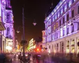 Nocą ulica Piotrkowska w Łodzi rozświetlona fioletowymi i zielonymi światłami. Widać ludzi spacerujących i siedzących w ogródkach kawiarnianych. Co ciekawego w Łodzi? Ta iluminacja!
