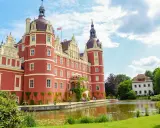 Czerwony zamek z wieżami, otoczony zielenią i stawem, stanowi serce parku mużakowskiego.
