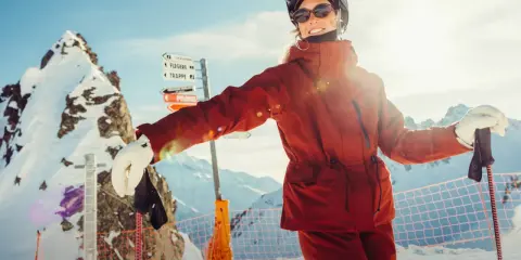 Narciarka w pomarańczowej kurtce Helly Hansen, z nartami i kijami, na tle ośnieżonych gór.