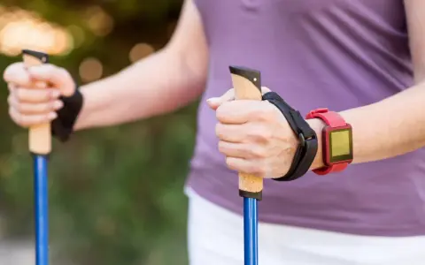 Dłonie trzymające kijki do nordic walking, z czerwonym zegarkiem na nadgarstku. To świetny sposób, jak chodzić z kijkami, by wzmocnić ciało.