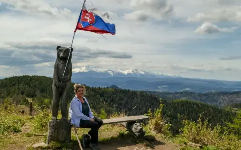Kobieta siedzi na ławce pod słowacką flagą na wietrznym wierchu. W tle rozciągają się góry i lasy.