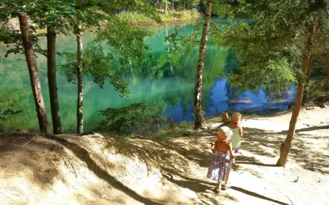 Dwie dziewczynki w słomkowych kapeluszach idą ścieżką nad turkusowe jezioro. W tle piękne góry i las.