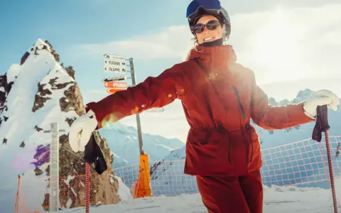 Narciarka w pomarańczowej kurtce Helly Hansen, z nartami i kijami, na tle ośnieżonych gór.