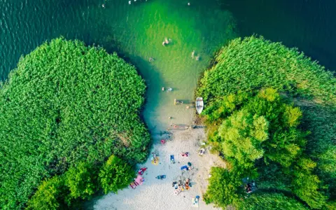 Letnie słońce ogrzewa plażowiczów nad jednym z najpiękniejszych jezior w Polsce. Woda kusi kąpiących się, a łódka czeka przy brzegu.