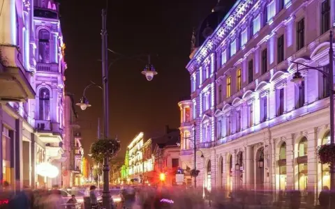 Nocą ulica Piotrkowska w Łodzi rozświetlona fioletowymi i zielonymi światłami. Widać ludzi spacerujących i siedzących w ogródkach kawiarnianych. Co ciekawego w Łodzi? Ta iluminacja!