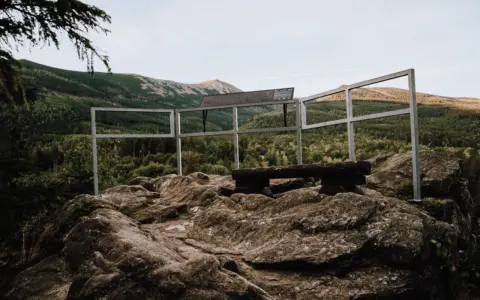 Kamienna platforma widokowa na szczycie, z ławką i balustradą, oferuje panoramę gór porośniętych lasem. To idealne miejsce na odpoczynek i podziwianie krajobrazu, niczym z legend o kruczych skałach.