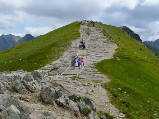 Tłum ludzi wspina się kamiennymi schodami na Kasprowy Wierch. Widać zielone zbocza i skaliste szczyty Tatr.