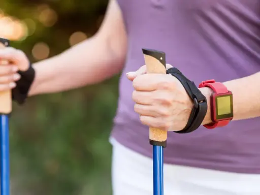 Dłonie trzymające kijki do nordic walking, z czerwonym zegarkiem na nadgarstku. To świetny sposób, jak chodzić z kijkami, by wzmocnić ciało.
