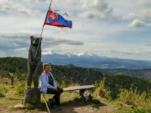 Kobieta siedzi na ławce pod słowacką flagą na wietrznym wierchu. W tle rozciągają się góry i lasy.