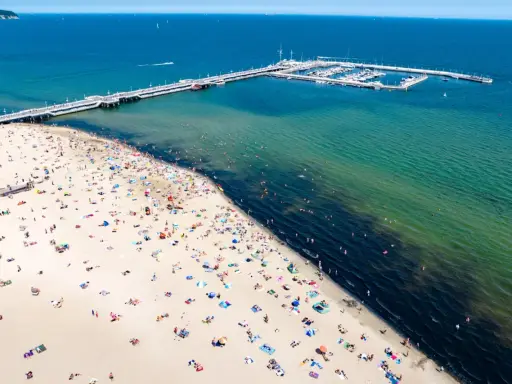 Tłum ludzi wypoczywa na piaszczystej plaży w Sopocie. W oddali widać molo i przystań jachtową.