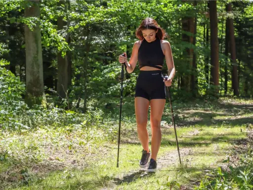 Młoda kobieta w czarnym sportowym stroju uprawia sport z kijami w lesie.