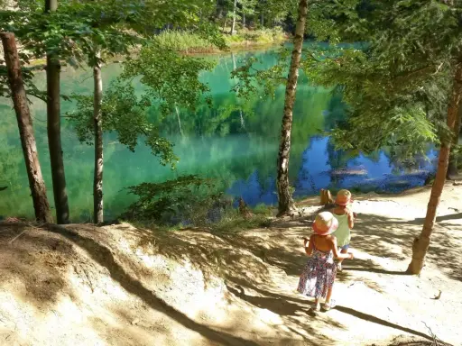 Dwie dziewczynki w słomkowych kapeluszach idą ścieżką nad turkusowe jezioro. W tle piękne góry i las.