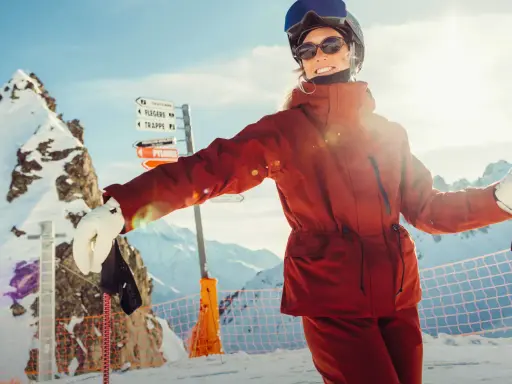 Narciarka w pomarańczowej kurtce Helly Hansen, z nartami i kijami, na tle ośnieżonych gór.