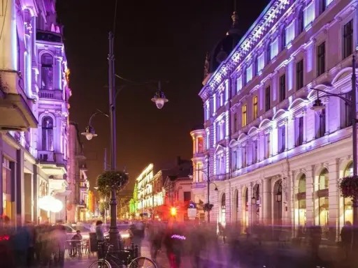 Nocą ulica Piotrkowska w Łodzi rozświetlona fioletowymi i zielonymi światłami. Widać ludzi spacerujących i siedzących w ogródkach kawiarnianych. Co ciekawego w Łodzi? Ta iluminacja!