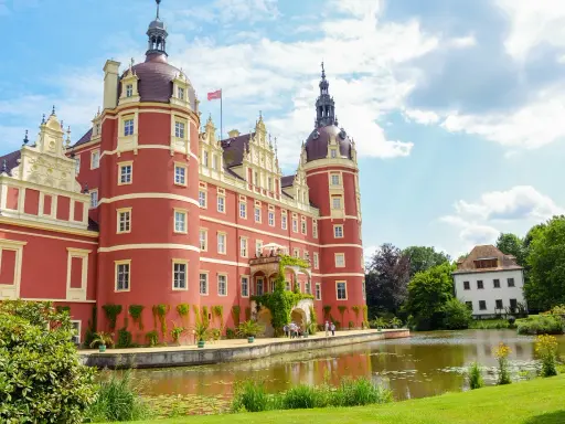 Czerwony zamek z wieżami, otoczony zielenią i stawem, stanowi serce parku mużakowskiego.