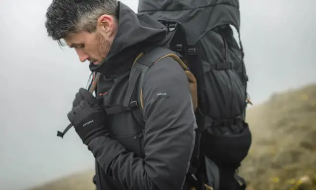 Mężczyzna w ciemnej kurtce softshell i rękawiczkach, z dużym plecakiem, przygotowuje się do wędrówki. Czy softshell jest nieprzemakalny? Ta kurtka wygląda na gotową na deszcz.