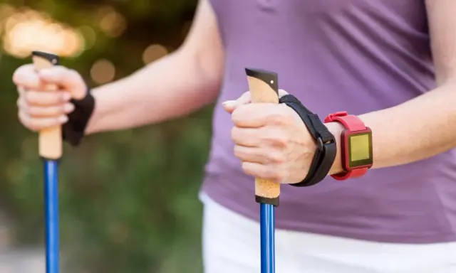 Dłonie trzymające kijki do nordic walking, z czerwonym zegarkiem na nadgarstku. To świetny sposób, jak chodzić z kijkami, by wzmocnić ciało.