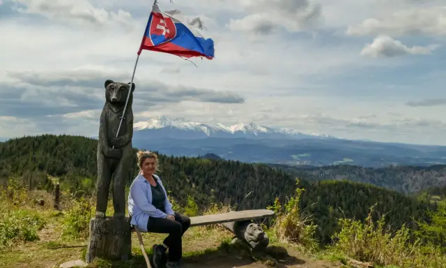 Kobieta siedzi na ławce pod słowacką flagą na wietrznym wierchu. W tle rozciągają się góry i lasy.