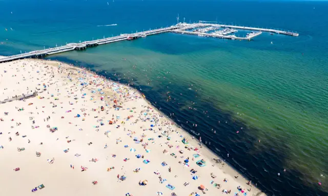 Tłum ludzi wypoczywa na piaszczystej plaży w Sopocie. W oddali widać molo i przystań jachtową.