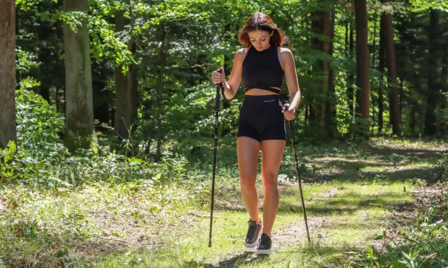 Młoda kobieta w czarnym sportowym stroju uprawia sport z kijami w lesie.