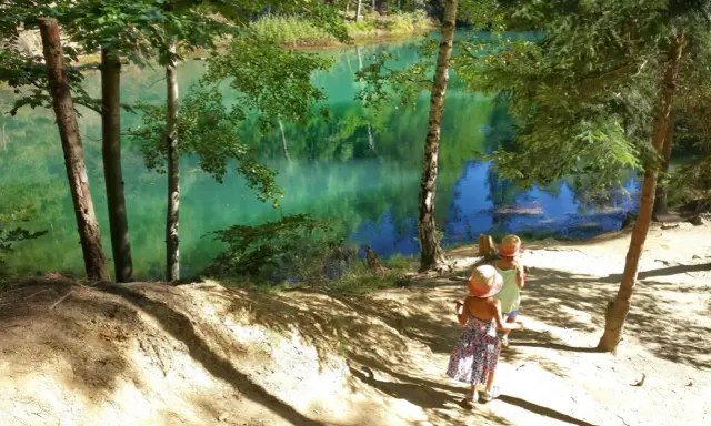 Dwie dziewczynki w słomkowych kapeluszach idą ścieżką nad turkusowe jezioro. W tle piękne góry i las.
