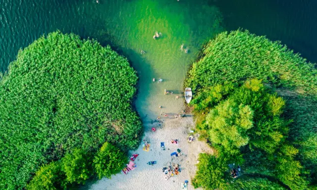 Letnie słońce ogrzewa plażowiczów nad jednym z najpiękniejszych jezior w Polsce. Woda kusi kąpiących się, a łódka czeka przy brzegu.