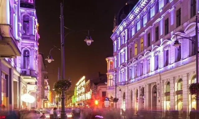 Nocą ulica Piotrkowska w Łodzi rozświetlona fioletowymi i zielonymi światłami. Widać ludzi spacerujących i siedzących w ogródkach kawiarnianych. Co ciekawego w Łodzi? Ta iluminacja!