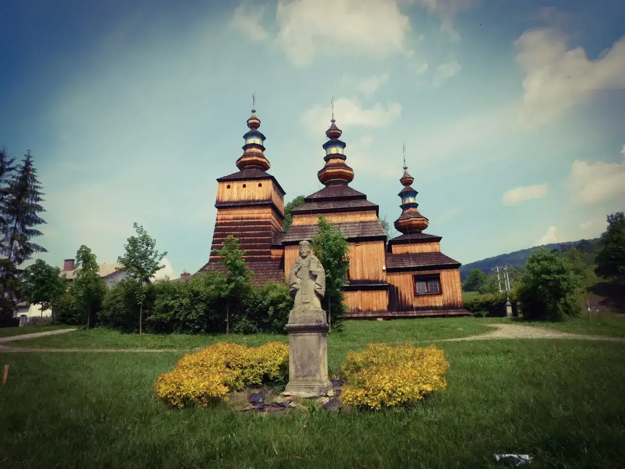 Drewniana cerkiew z trzema kopułami, kamienny pomnik i żółte krzewy. Beskid Niski atrakcje, które warto odkryć.