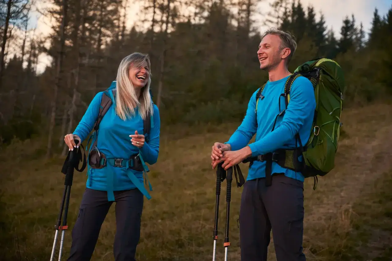 Para idąca przez las, ubrana na cebulkę w turkusowe bluzy i ciemne spodnie, z plecakami i kijkami trekkingowymi.