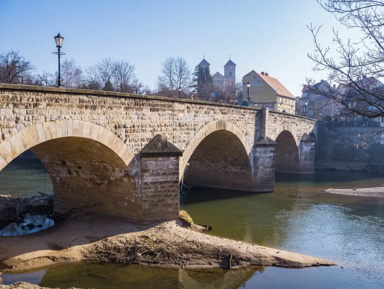 Kamienny most z arkadami nad rzeką w Bardo Śląskim. W tle kościół i zabudowania miejskie.