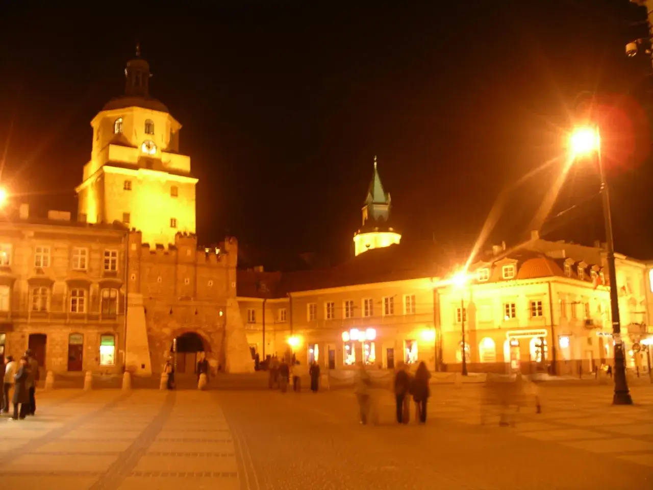Nocne lublin atrakcje: Brama Krakowska i Rynek rozświetlone latarniami, z sylwetkami przechodniów.
