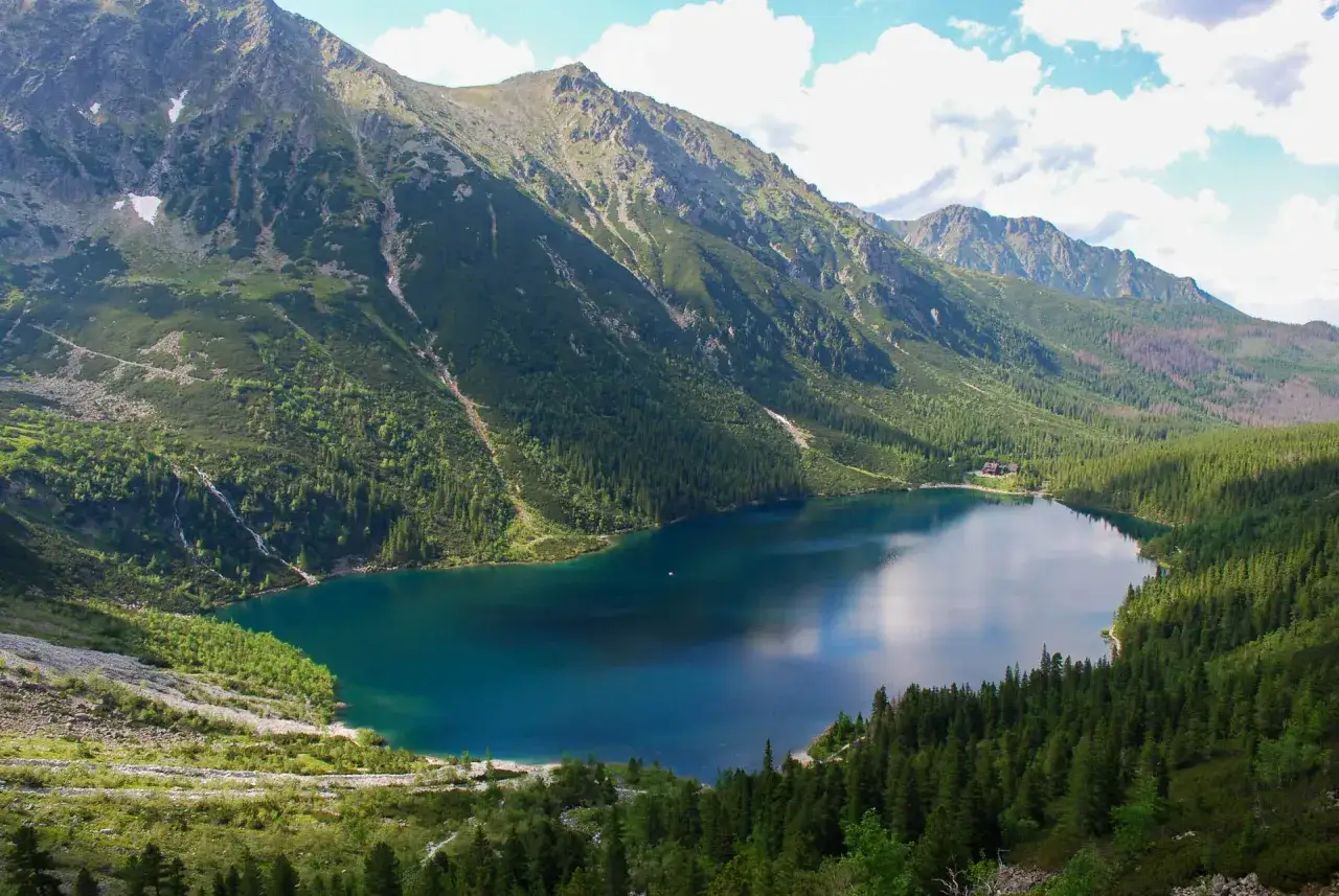 Malownicze Tatry z jeziorem Morskie Oko. W oddali widać schronisko, a zbocza g&oacute;r porasta gęsty las.
