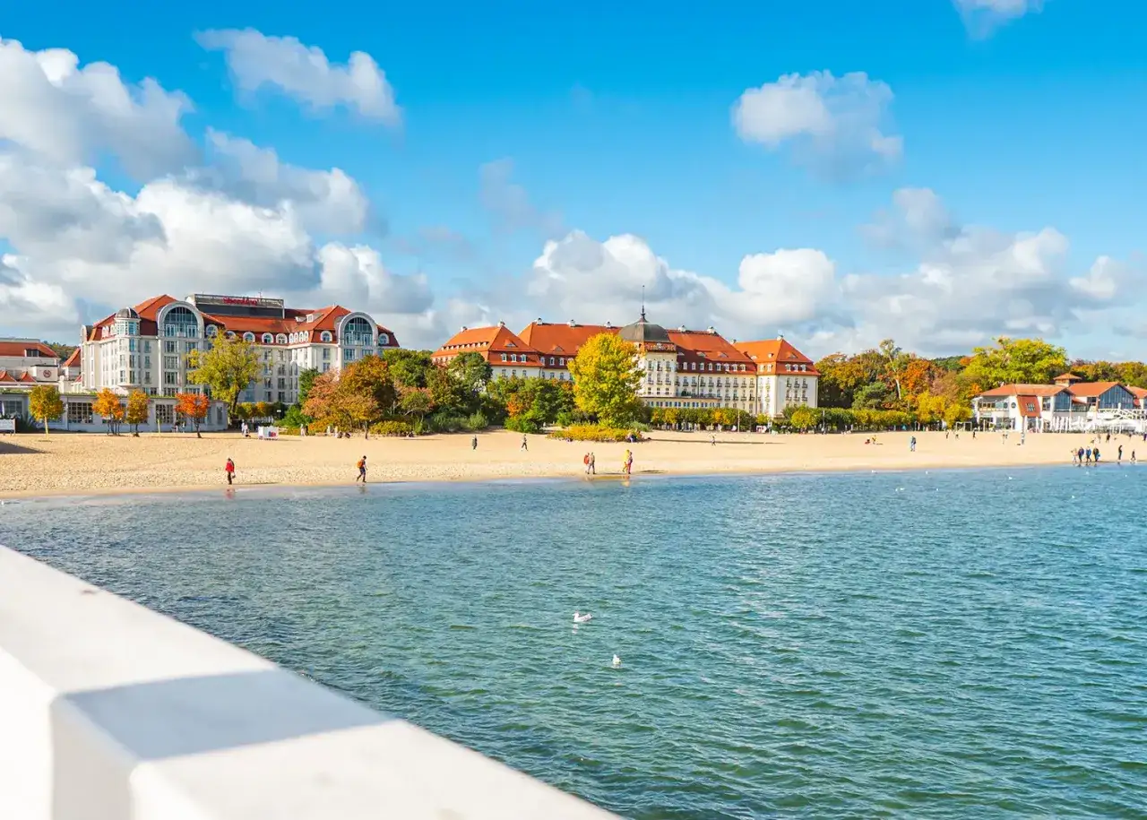 Piękna plaża w Sopocie z widokiem na zabytkowe budynki i morze. Kilka osób spaceruje po piasku, ciesząc się jesiennym dniem.