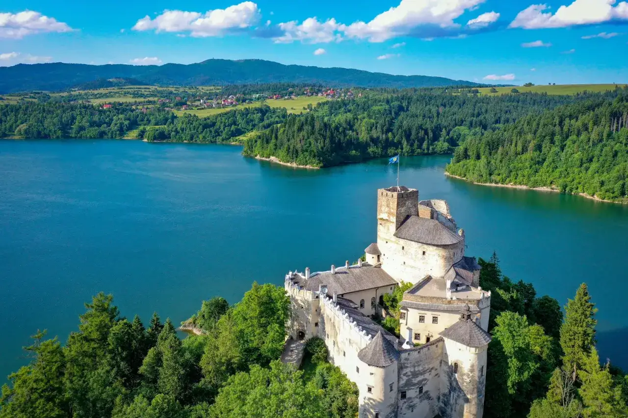 Zamek w Niedzicy góruje nad turkusowym jeziorem czorsztyńskim, otoczony zielonymi lasami i pagórkami.