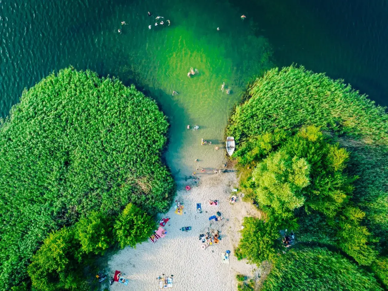 Letnie słońce, plaża i kąpiel w jednym z najpiękniejszych jezior w Polsce. Ludzie kąpią się, odpoczywają na piasku, a łódka czeka przy brzegu.