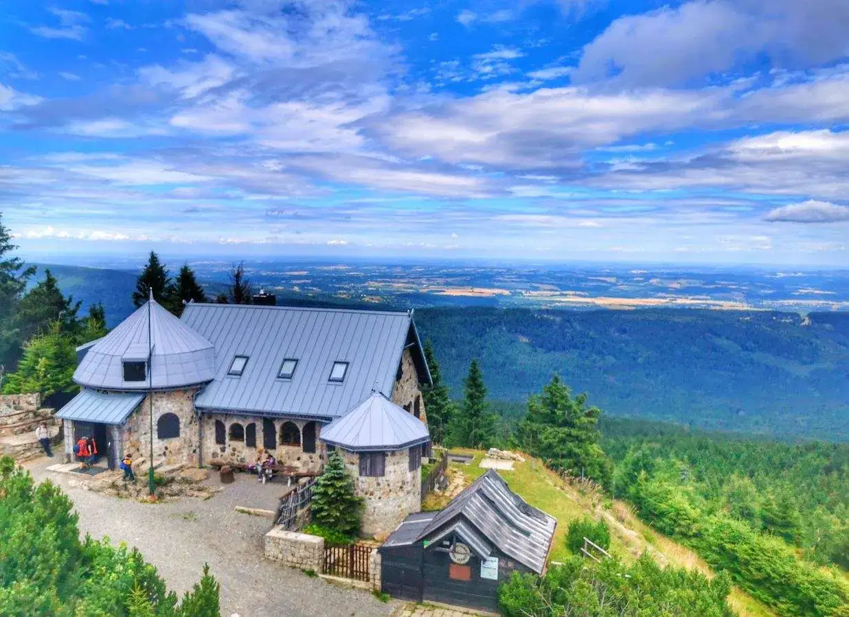 Kamienny budynek z szarym dachem na tle rozległej panoramy G&oacute;r Izerskich. W oddali widać pola i lasy.