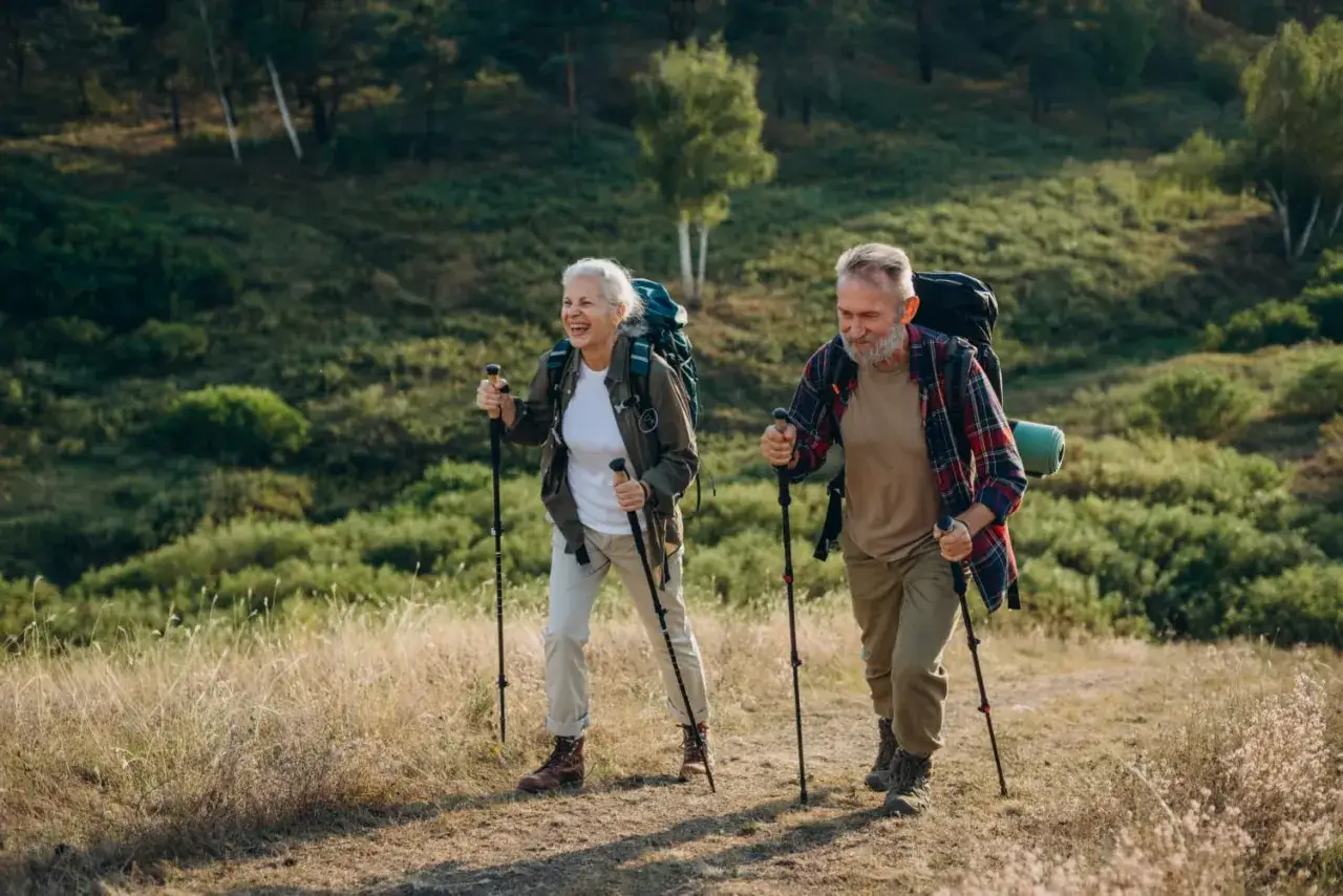 Uśmiechnięta para seniorów z plecakami przemierza górski szlak, ucząc się, jak chodzić z kijkami.